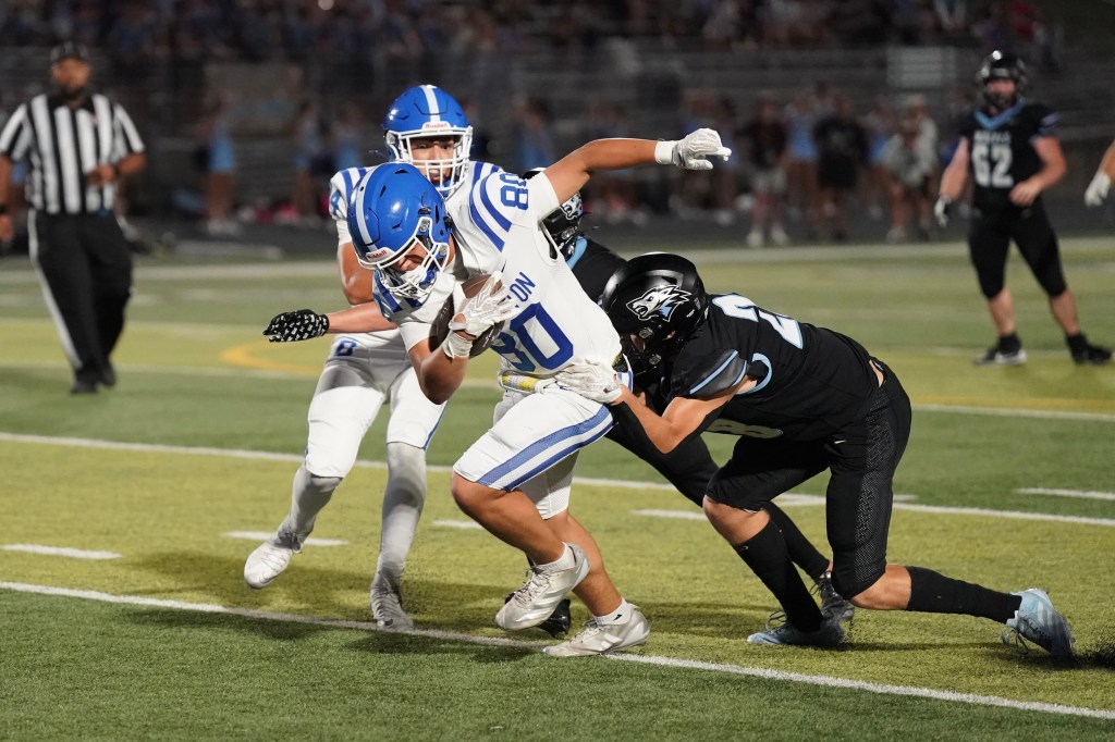 A football player in a white jersey with blue accents is being tackled by an opponent in a black jersey. Two teammates in blue jerseys are nearby, and a referee is in the background.