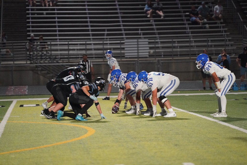 Two football teams lined up on the field during a night game, one team in blue and white uniforms and the other in black uniforms, preparing for a play.