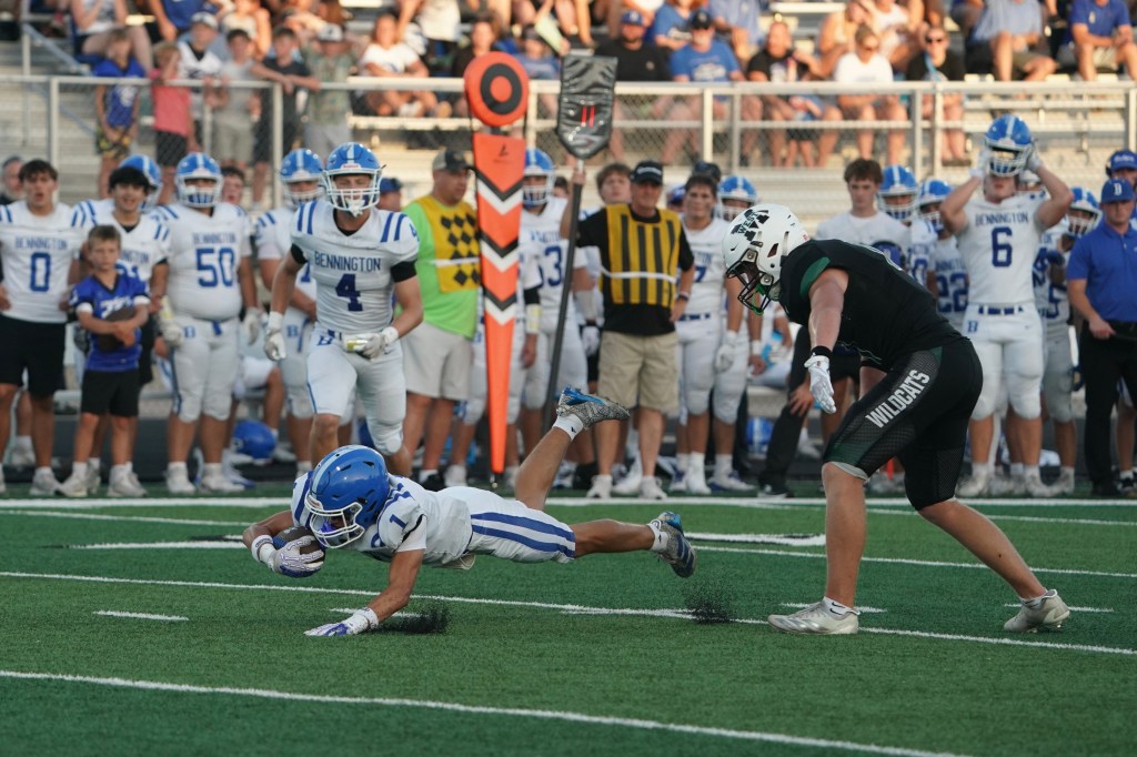 A football player from Bennington dives to catch the ball while being pursued by an opposing player in a black jersey. Spectators in the background are cheering.