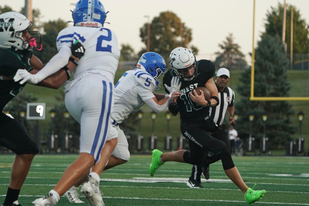 A high school football player in a black uniform running with the ball, while another player in a blue uniform attempts to tackle him. The scene takes place on a football field with goalposts in the background.
