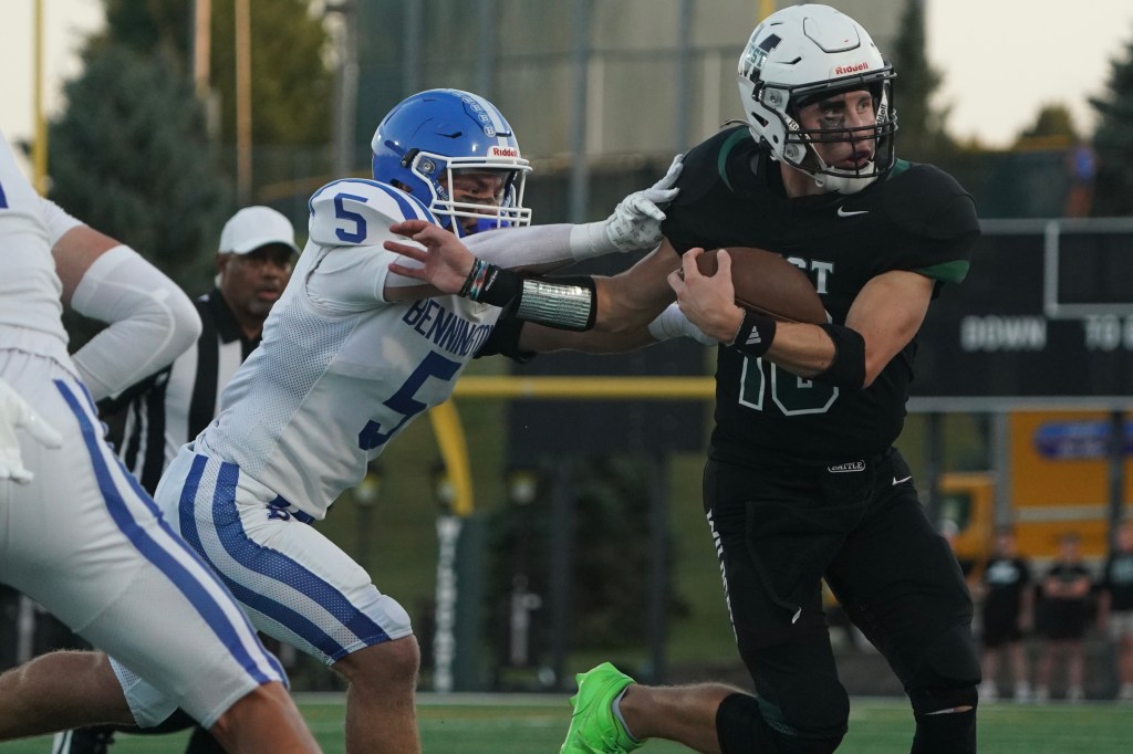 A high school football player in black jersey number 10 evades a defender in a white jersey number 5 while carrying the ball during a game.