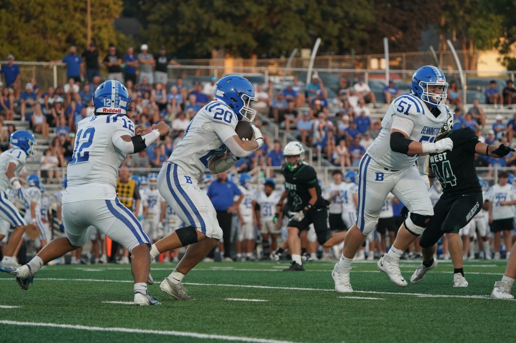 A high school football game in progress, featuring players in white and blue uniforms on offense, with a crowd of spectators in the background.