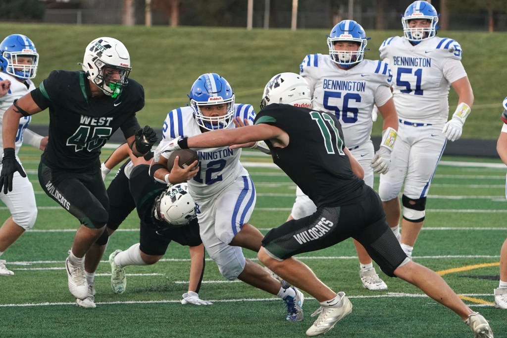 A high school football game scene showing players in action. One player in a blue jersey, number 12, is being tackled by two defenders in black jerseys, while other players from both teams are in the background.