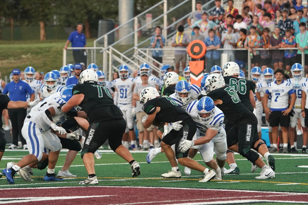High school football players compete on the field, with a mix of black and white uniforms, while spectators watch from the stands.