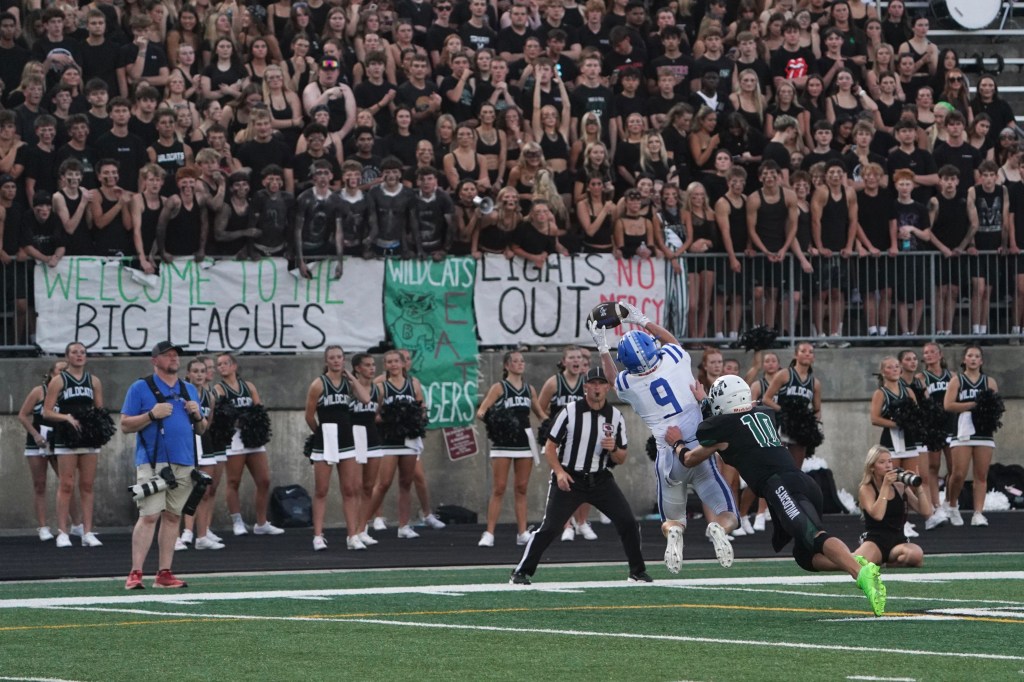A high school football game scene with a player in a blue and white uniform catching a pass while being defended by a player in a black uniform. In the background, a large crowd wearing black cheers with banners, and cheerleaders in black and white are on the sidelines.