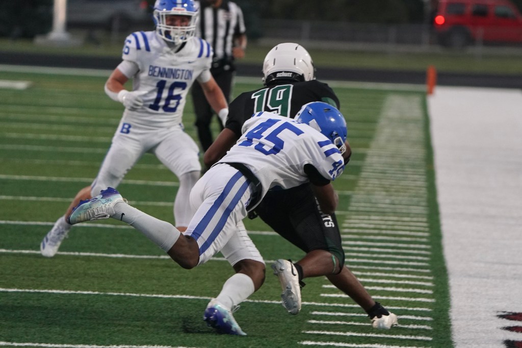 A football player in a blue jersey tackles an opponent in a black jersey during a game on a green turf field.