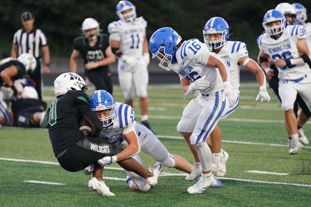 A high school football game with players in blue and white uniforms from Bennington tackling a player in black from the Wildcats team. The focus is on two Bennington players; one is about to tackle the opposing player.