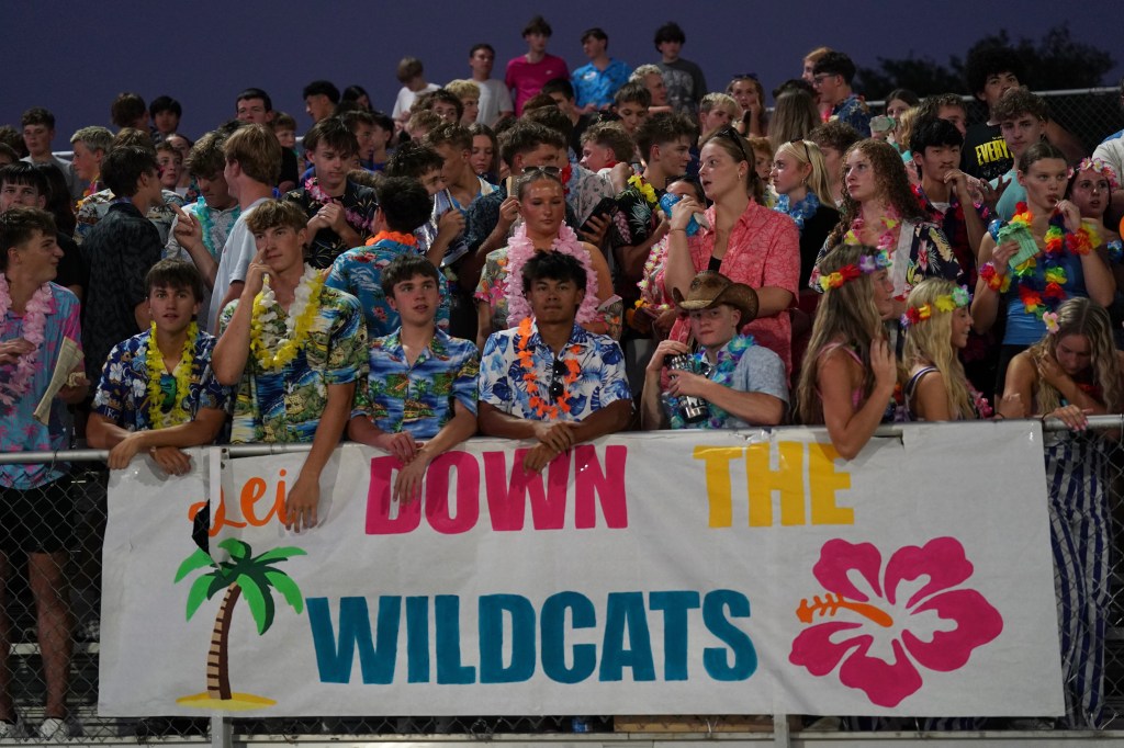 A crowd of high school students dressed in colorful Hawaiian shirts and leis, gathered at a sporting event, holding a banner that reads 'Let Down the Wildcats'.