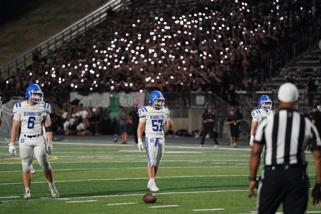 A football field at night with Bennington team players in white and blue uniforms, preparing for a play. A large crowd with lights in the background is cheering.
