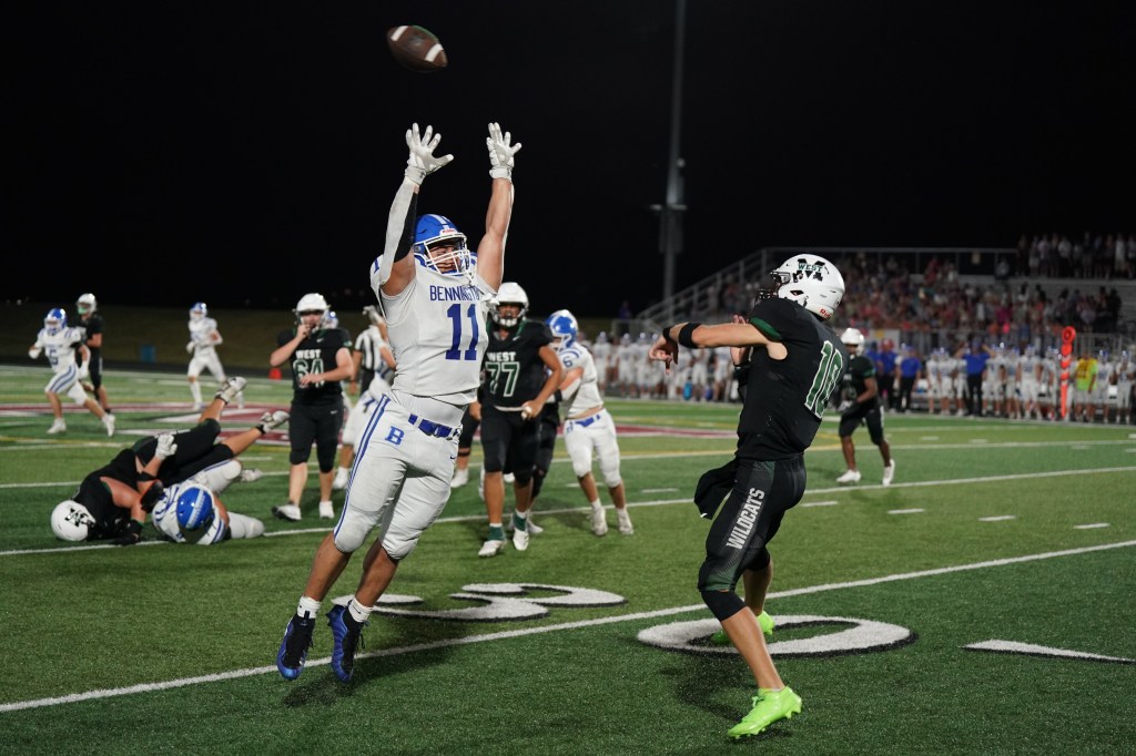 A football player in a white uniform leaps to catch a ball during a night game, while another player in a black uniform throws the ball.