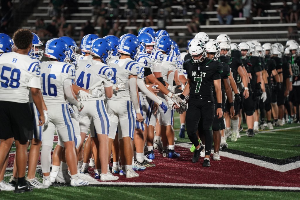 Two high school football teams shaking hands on the field after a game, one team in blue and white uniforms and the other in black uniforms.