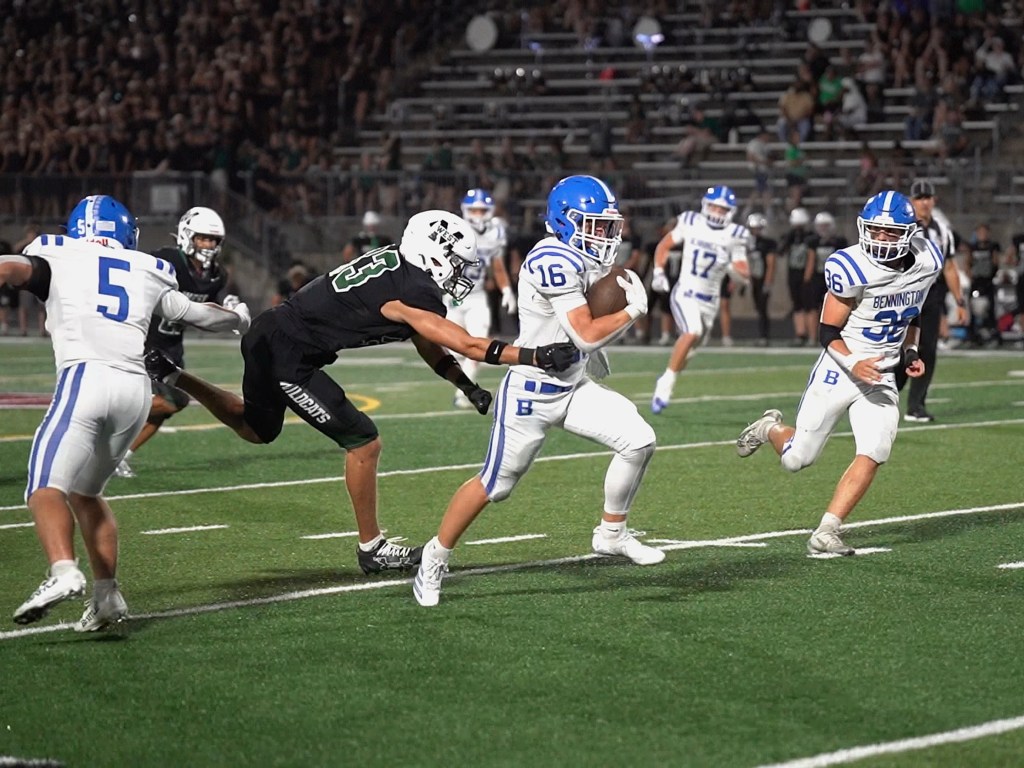 A high school football game in progress, with players wearing white and blue uniforms and a player in a black uniform attempting to tackle the player with the ball.
