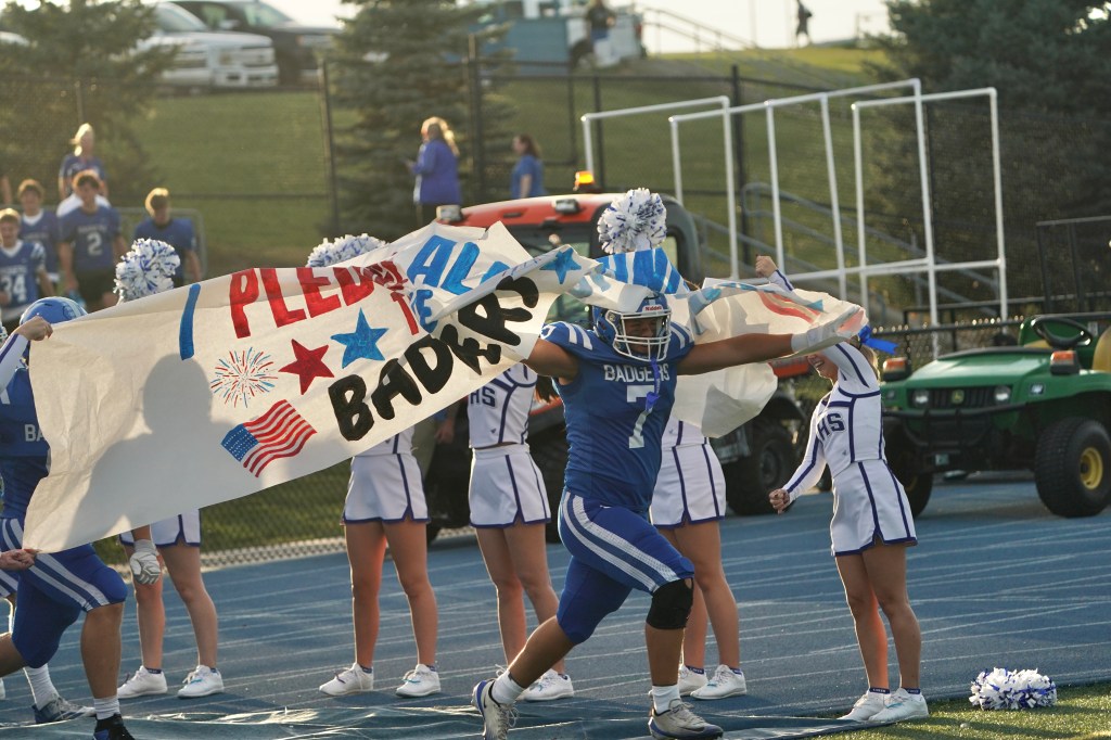 A football player in a blue jersey runs through a banner that reads 'I Pledge Allegiance' with patriotic decorations, while cheerleaders in uniforms cheer in the background at a sports event.