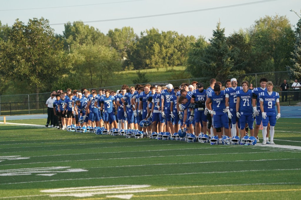 A line of football players in blue jerseys standing on the field with their helmets lined up in front of them, during a team event.