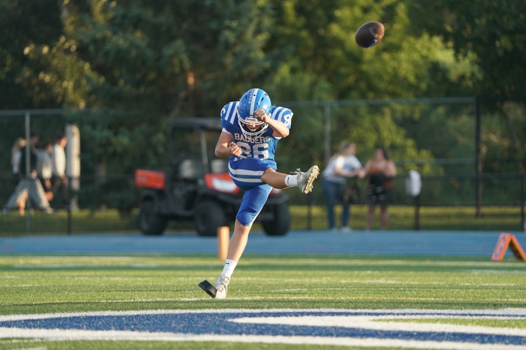A high school football player in a blue uniform prepares to kick a football during a game, with spectators in the background.