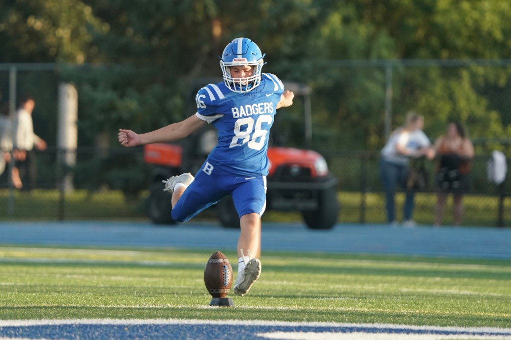 A football player in a blue jersey with the number 86 is preparing to kick a football on a field. The player stands on grass with a blurred background of spectators and equipment.