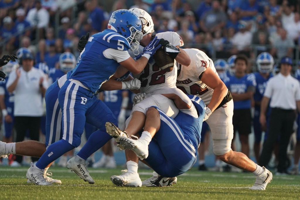 Two football players in blue jerseys tackle an opposing player in a white jersey on the field during a game.