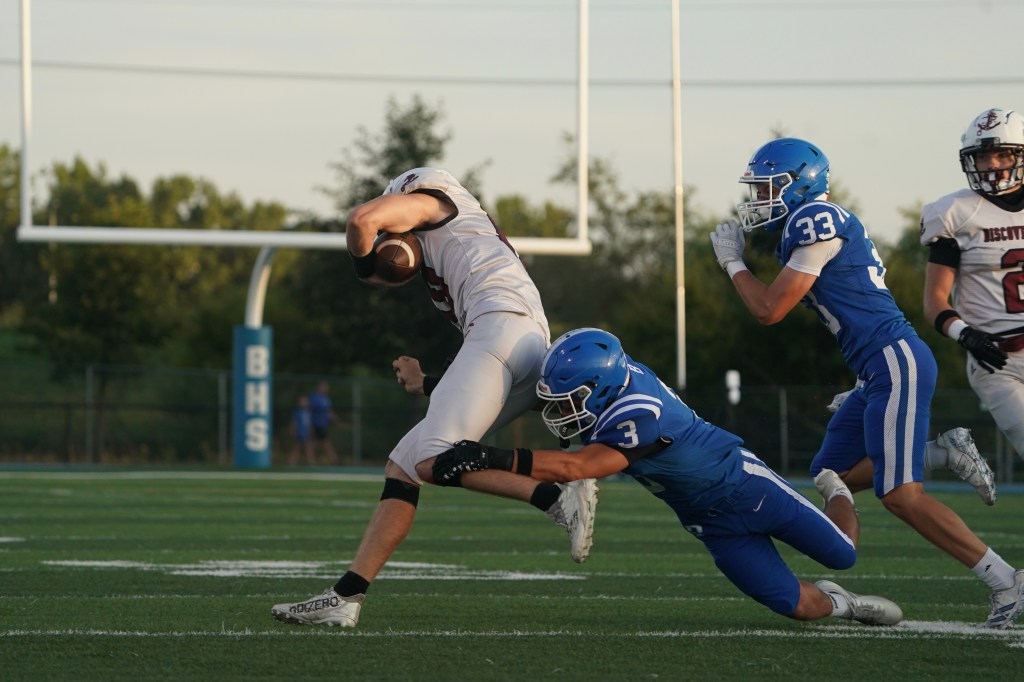 A high school football player in a white uniform is being tackled from behind by a player in a blue uniform, while another blue-uniformed player is nearby. The scene takes place on a grassy football field with goalposts in the background.