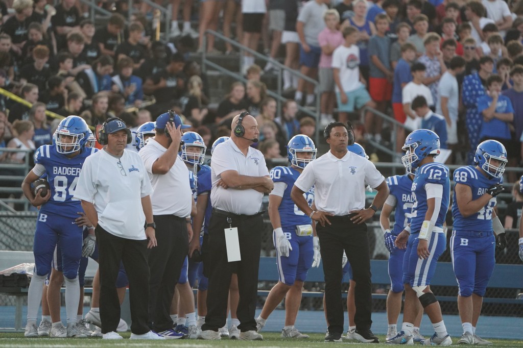 A group of football coaches and players in blue uniforms are gathered on the sidelines during a game, with a crowd of spectators visible in the background.