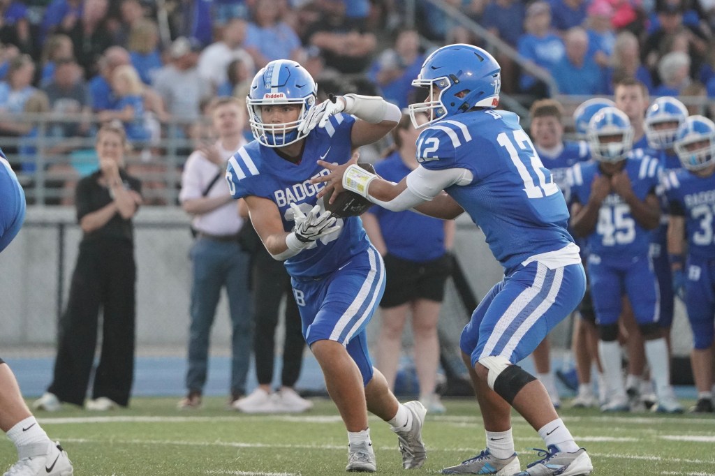 Two high school football players in blue uniforms are executing a play on the field, with one player handing off the football to the other while fans watch in the background.