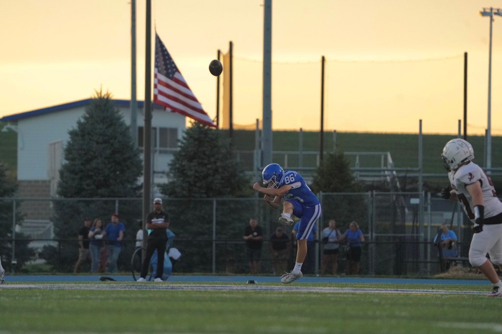 A football player in blue jersey number 86 kicks a ball during a game at sunset, with an American flag visible in the background.