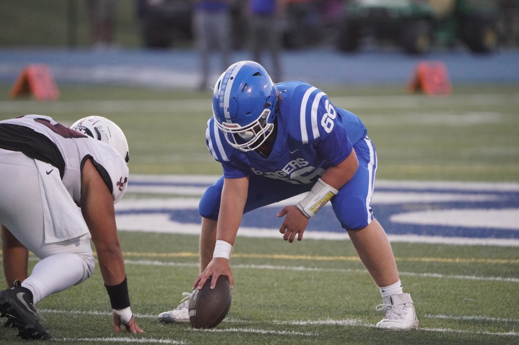 A football player in a blue jersey crouches on the field, preparing to snap the football to an opposing player in a black and white uniform, with players in the background.