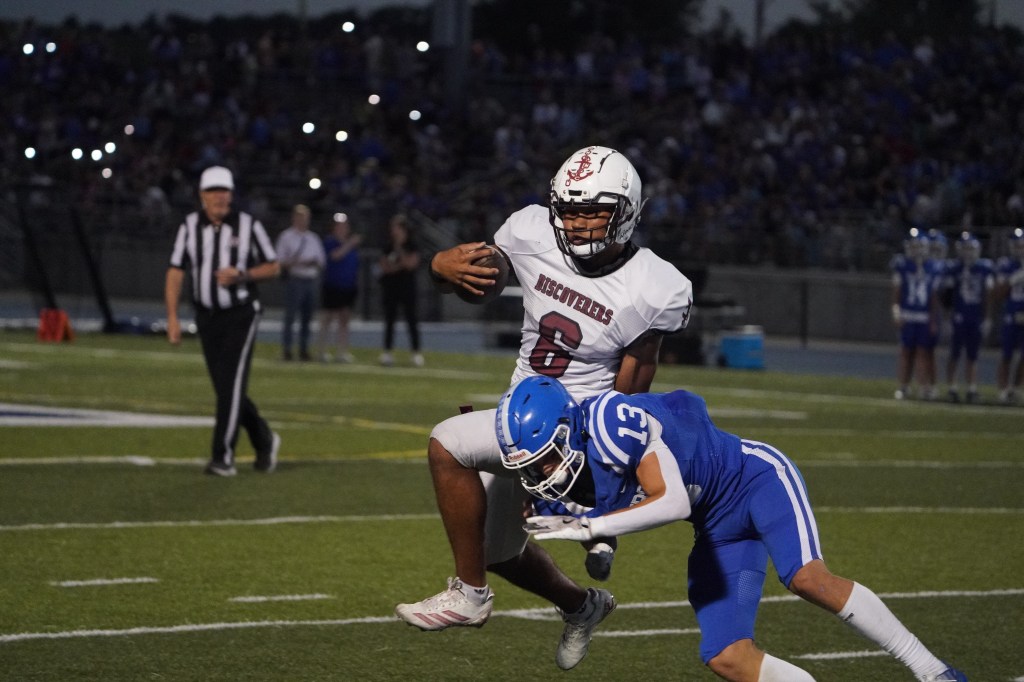 A football player wearing a white jersey with '6' running with the ball while being tackled by an opponent in a blue jersey with '13' during a night game.