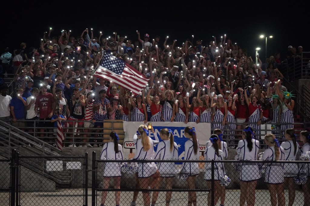 A large crowd at a nighttime event, many holding up their phone lights, waving an American flag. The crowd is dressed in patriotic colors, while cheerleaders in white outfits perform in front of them.