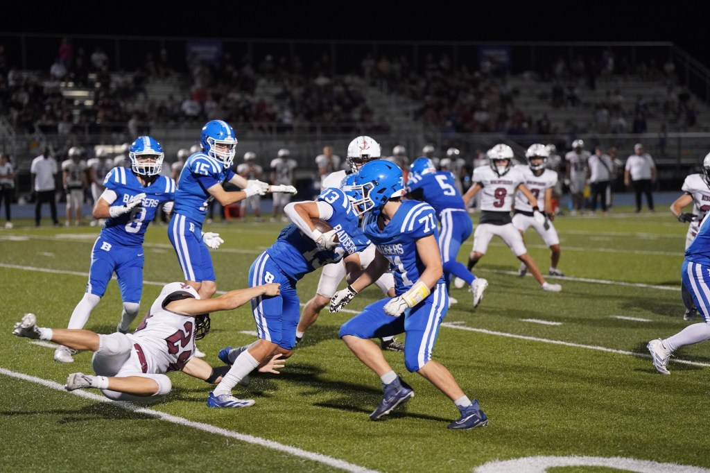 A high school football game in progress, featuring players in blue and white uniforms on the field. One player is evading a tackle while others are positioned in the background.