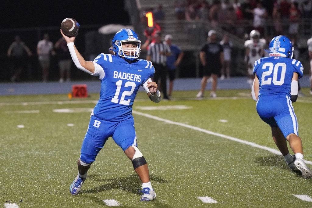 A high school football player in a blue jersey with 'BADGERS' and the number '12' throwing a football on a field during a night game.