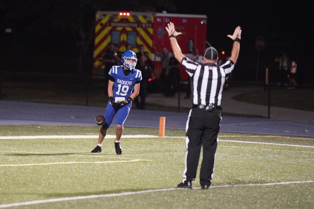A high school football player in a blue uniform with 'Badgers' on the front, wearing number 19, celebrating on the field under stadium lights. A referee in black and white stripes is signaling a play in the background.