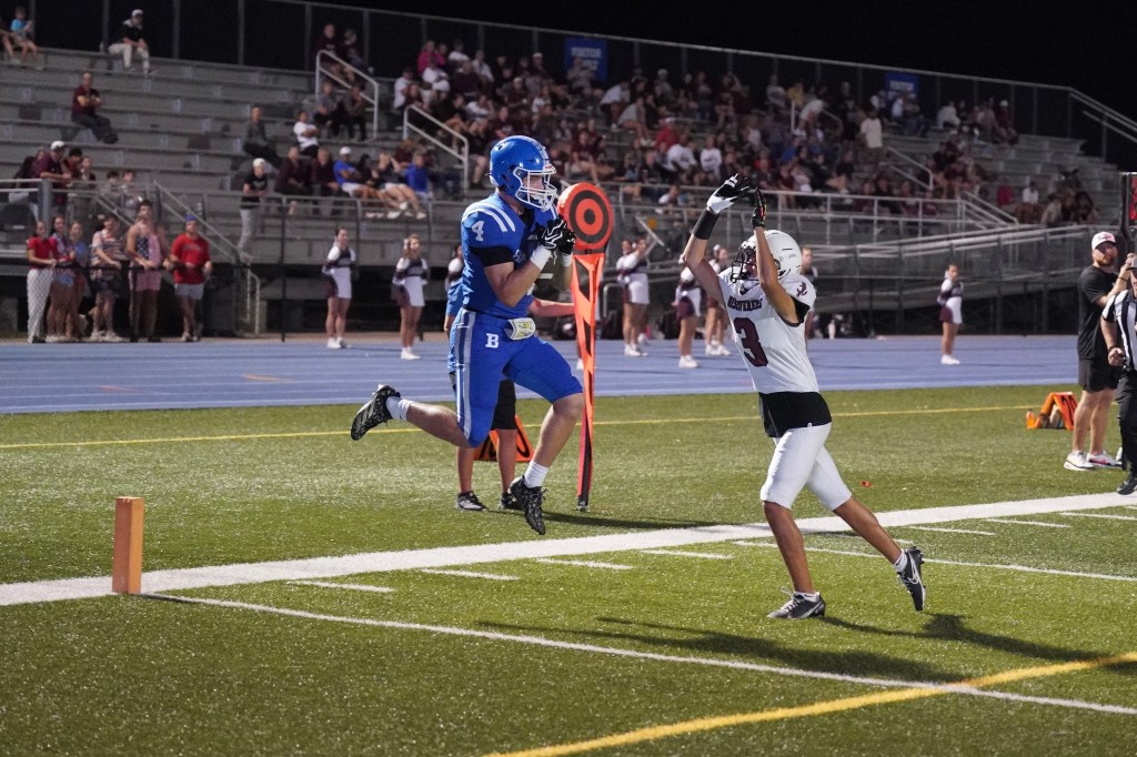 A blue jersey football player jumps to catch a pass in the end zone while a white jersey player tries to defend during a night game.