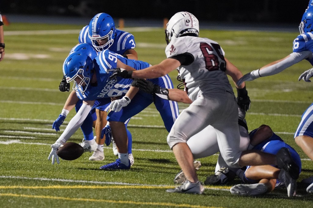 A high school football game scene showing players in blue and white uniforms attempting to recover a loose ball on the field, with some players diving toward the ball while others engage in a tackle.