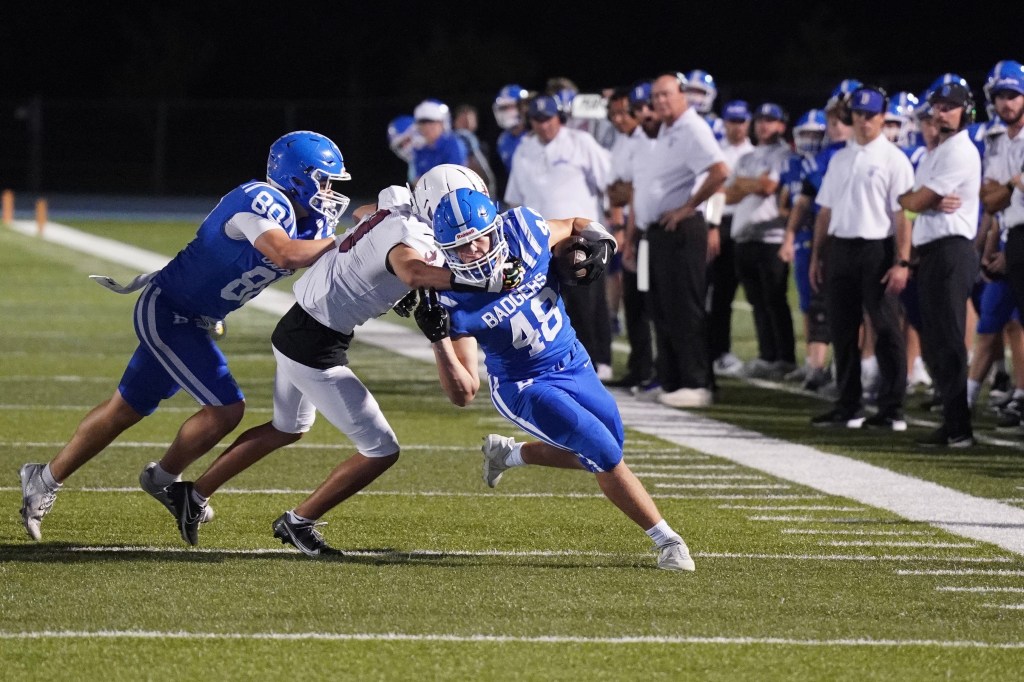 A blue jersey football player is being tackled by two opponents during a night game on a turf field.