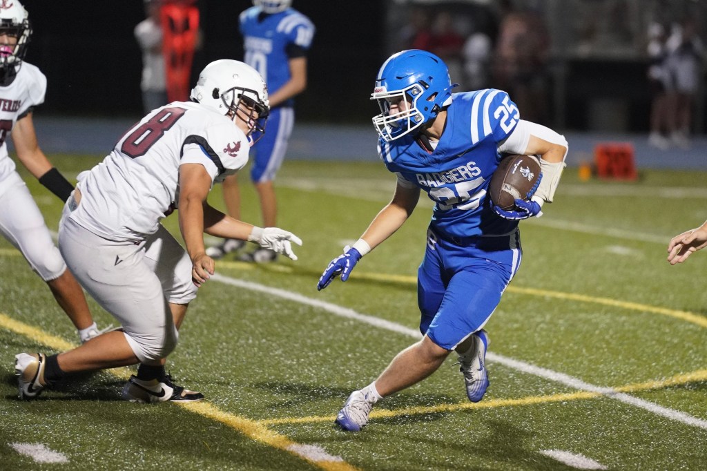 A football player in a blue jersey with the number 23 is running with the ball, while a player in a white jersey with the number 8 attempts to tackle him during a night game on a grass field.