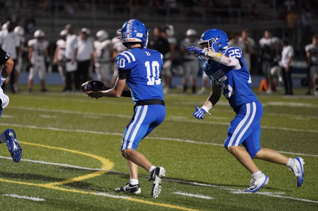 Two high school football players in blue uniforms, one quarterback in number 10 jersey preparing to pass the ball, and another player in number 25 jersey running alongside to receive.