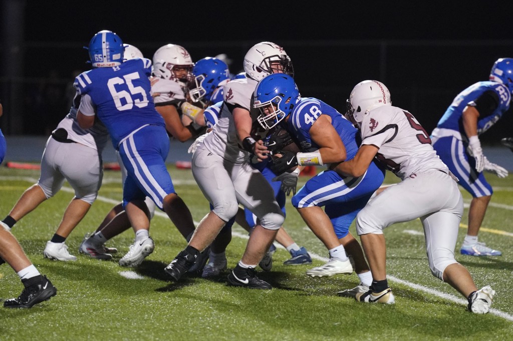 A high school football game in action, featuring players in blue and white uniforms engaged in a tackle on the field.