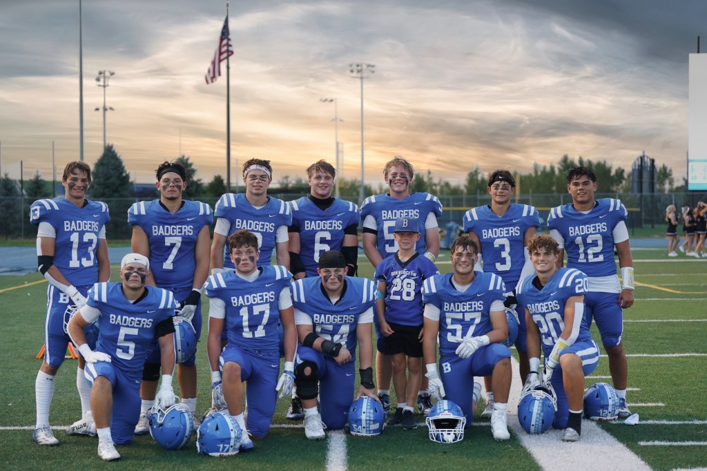 A group photo of a youth football team, the Badgers, posing on a field. The players are wearing blue jerseys with white stripes and helmets, with a sunset sky in the background.