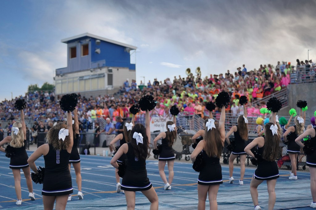 Cheerleaders performing in front of a large crowd at a sports event, with colorful attire in the stands and a blue sky overhead.