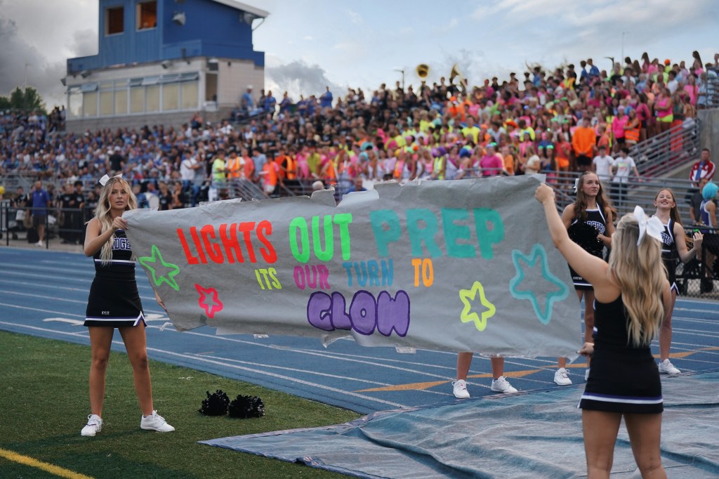 Cheerleaders holding a colorful banner that says 'LIGHTS OUT PREP ITS OUR TURN TO GLOW' during a school event, with a large crowd in the background.