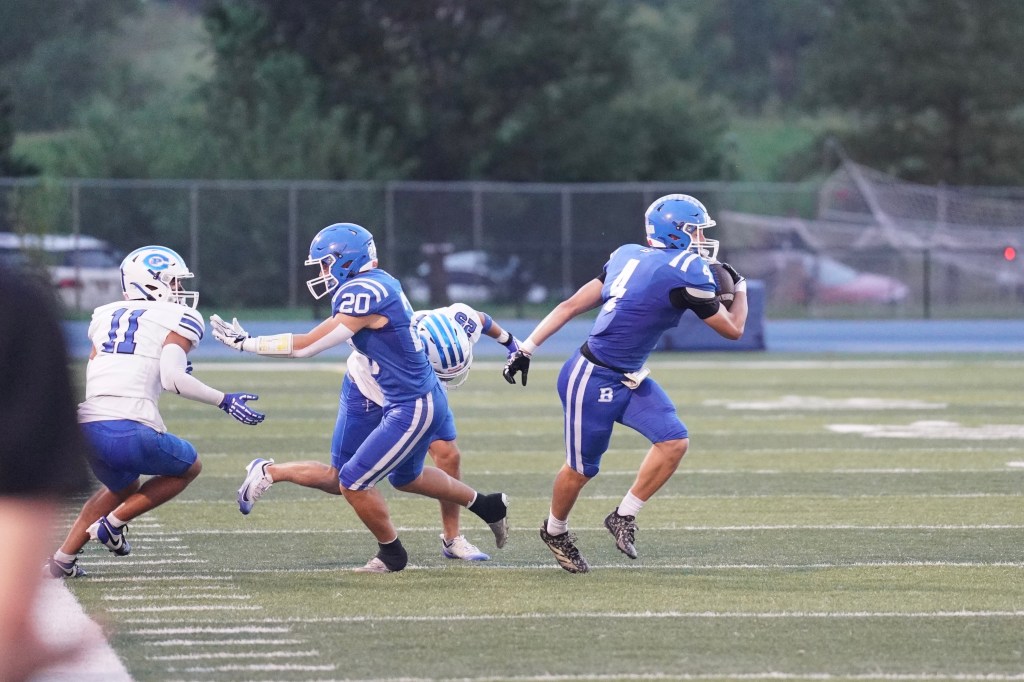 A football player in blue uniform, number 4, runs with the ball while evading a defender in white uniform, number 11. Another blue uniformed player, number 20, assists in blocking the opponent.