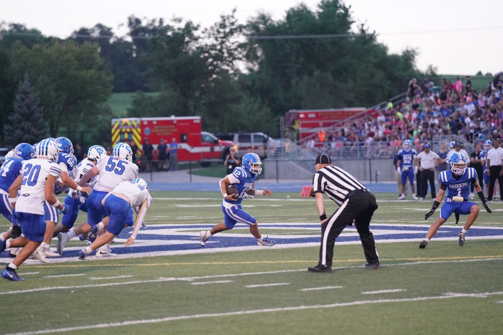 A high school football game in progress, featuring players in blue and white uniforms on a field. The action focuses on a player with the ball running towards the right, while a referee watches closely.
