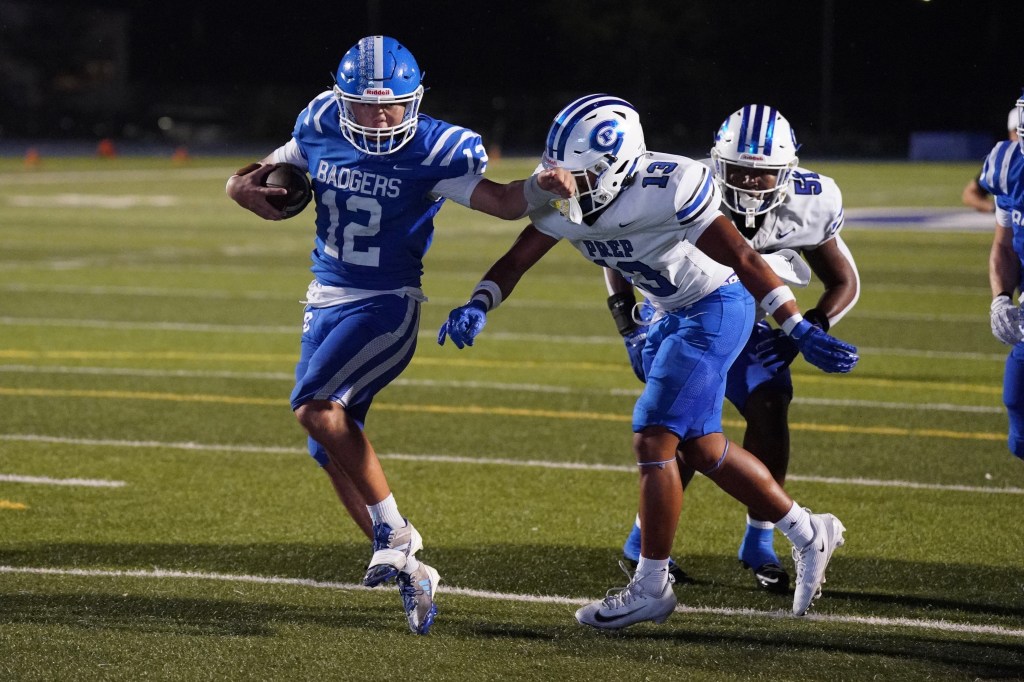 A high school football player wearing a blue jersey with the number 12 evades a tackle from an opponent wearing a white jersey with the number 13 during a game on a well-lit field.