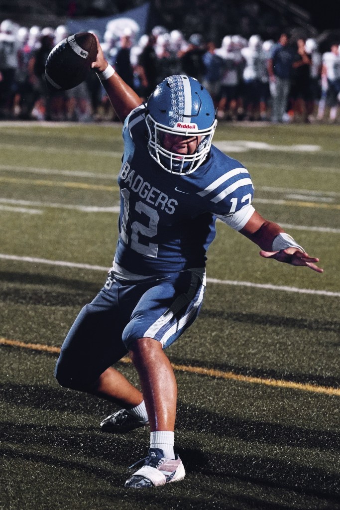 A football player wearing a blue jersey with 'Badgers' and the number 12 is in action, throwing a football on a field with spectators in the background.