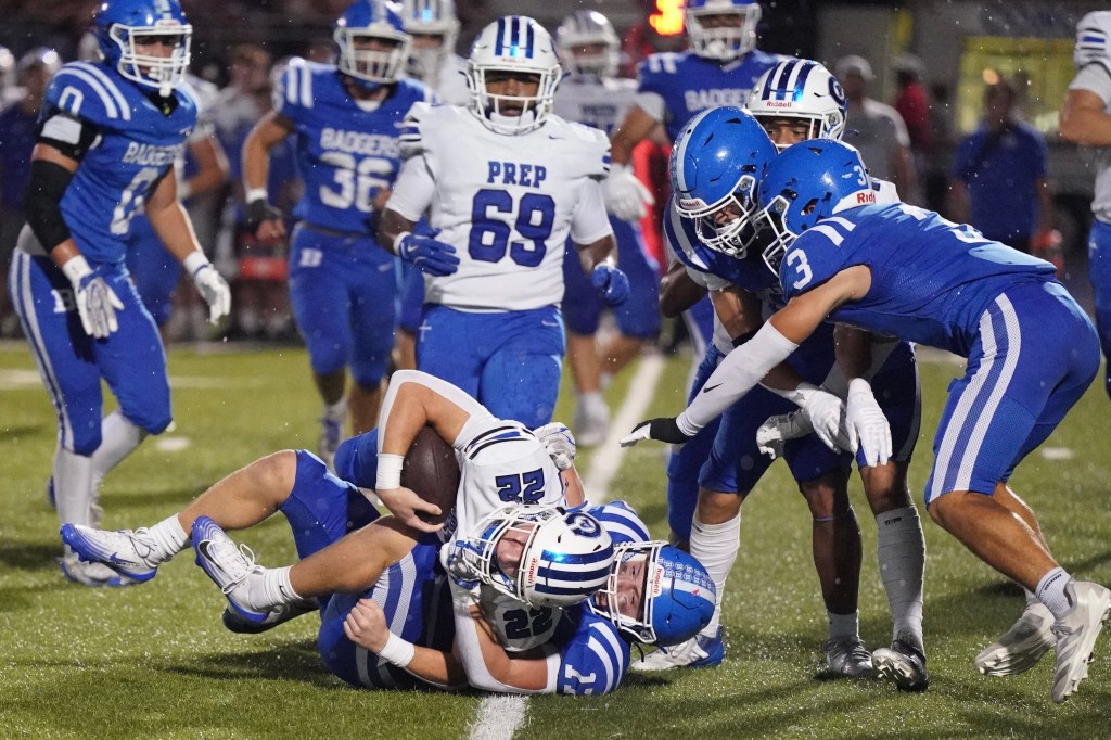 A football play where a player in blue is tackling another player in white on the field, surrounded by teammates and opponents.