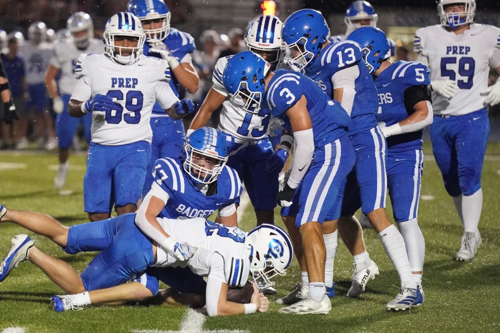 A group of high school football players in blue uniforms huddled around a player on the ground during a game, with players in white uniforms visible in the background.