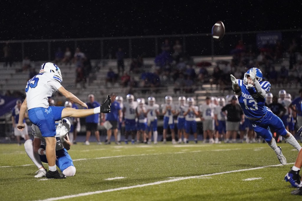 A football player in a white jersey, number 25, prepares to kick the ball while another player in blue, number 13, attempts to block the kick during a night game.