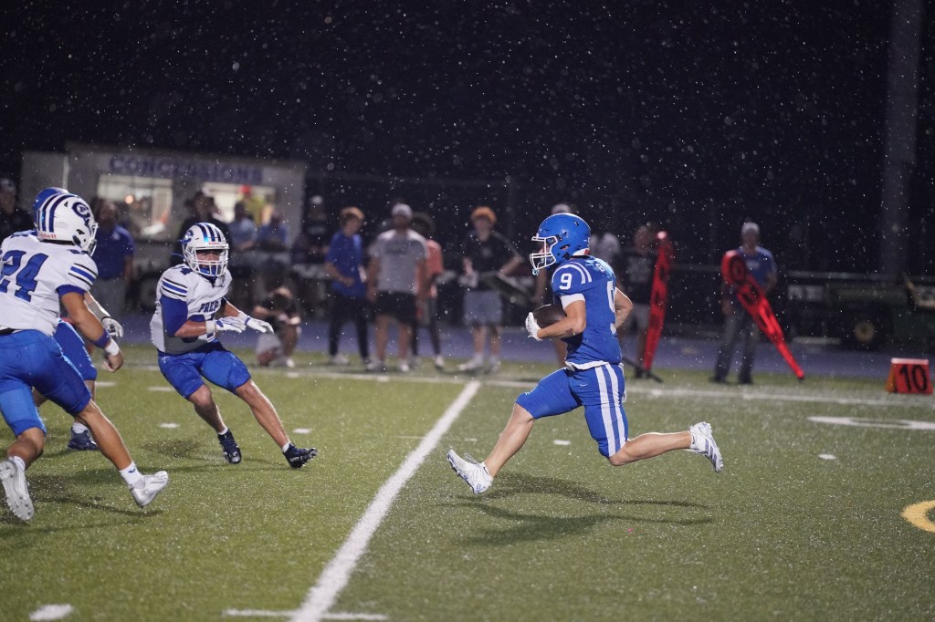 A football player in blue uniform runs with the ball on a wet field during a game in the rain, while two opposing players in white uniforms attempt to tackle him.