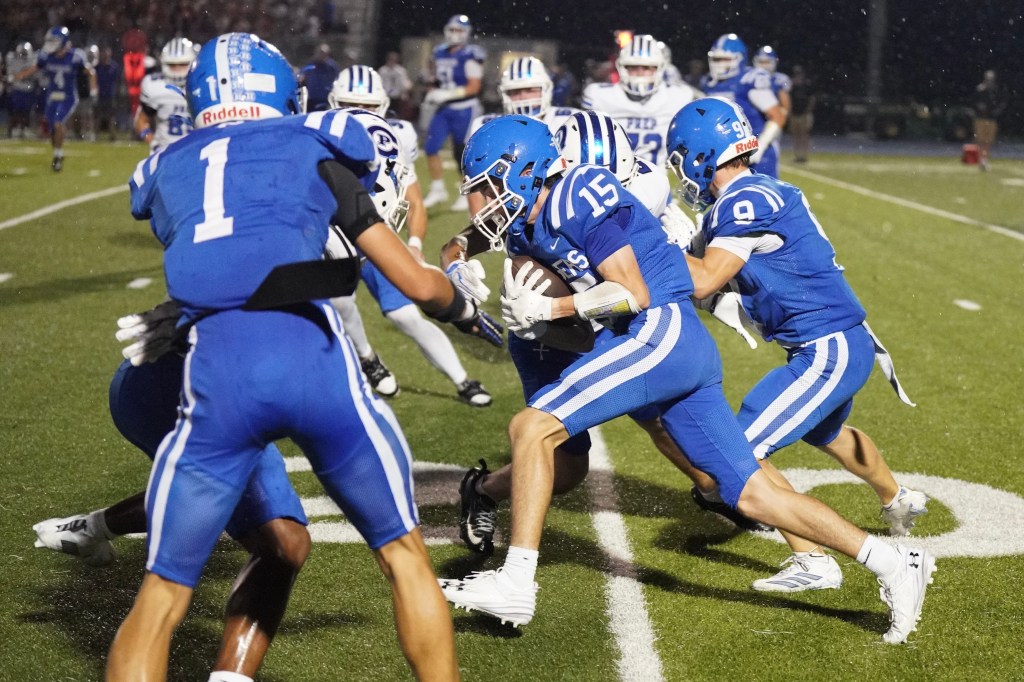 A high school football game scene showing players in blue uniforms tackling an opponent. The focus is on a player wearing the number 15, who is running with the ball, while teammates and opponents are engaged in the play.