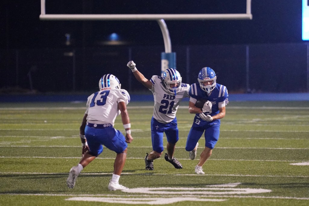 Two football players in blue uniforms and one in a white uniform engaged in action on the field at night.